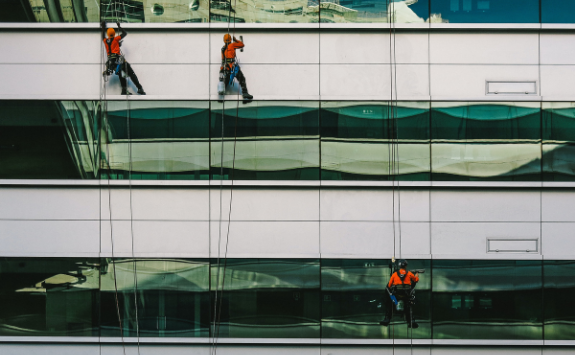 Workers clean the windows of a high-rise building. They are on safety ropes and wear safety gear.
